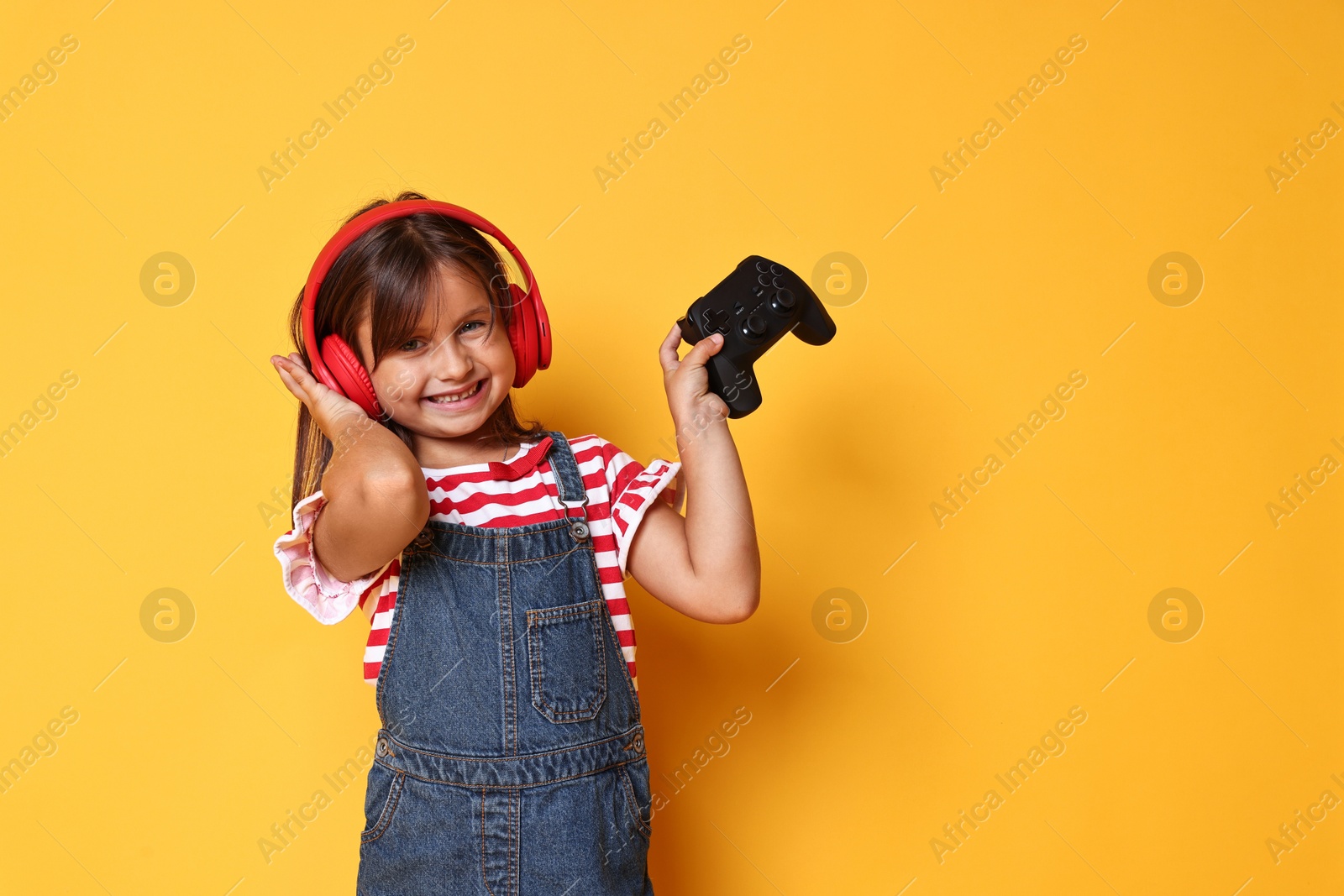 Photo of Smiling little girl in headphones with controller on orange background. Space for text