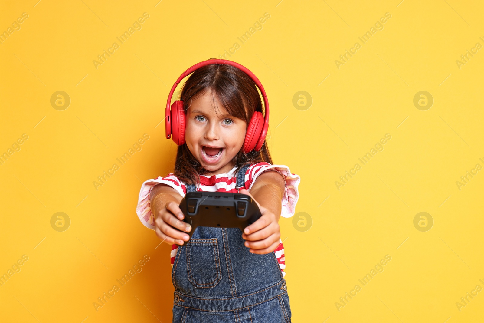 Photo of Cute little girl in headphones playing video game with controller on orange background