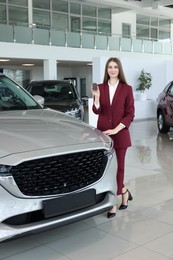 Photo of Happy saleswoman holding key near new silver car in salon