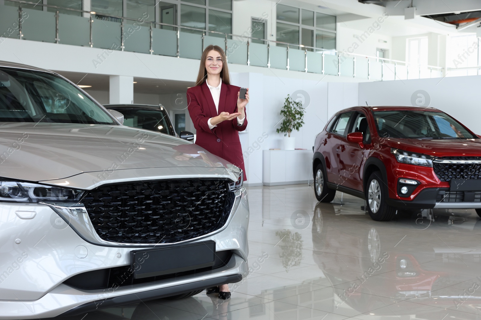 Photo of Happy saleswoman holding key near new silver car in salon