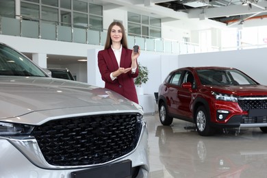 Photo of Happy saleswoman holding key near new silver car in salon