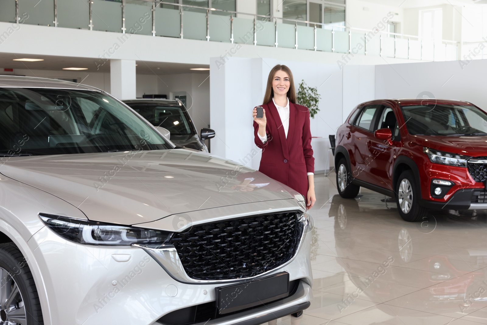 Photo of Happy saleswoman holding key near new silver car in salon