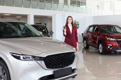 Photo of Happy saleswoman holding key near new silver car in salon