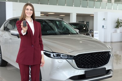 Photo of Happy saleswoman holding key near new silver car in salon