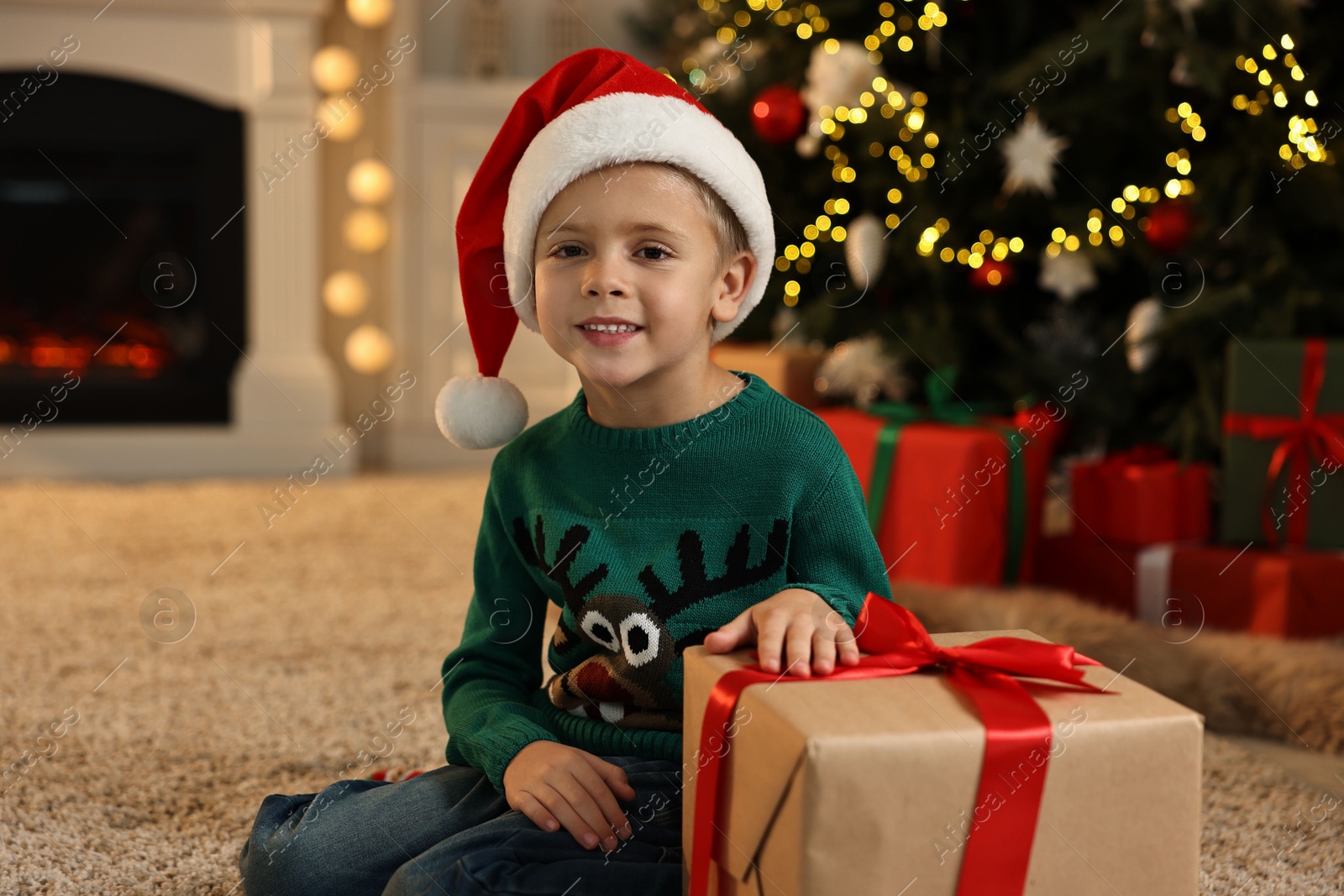 Photo of Cute little boy in Santa hat with Christmas gift indoors