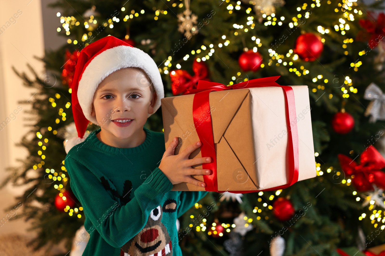 Photo of Cute little boy in Santa hat with Christmas gift indoors