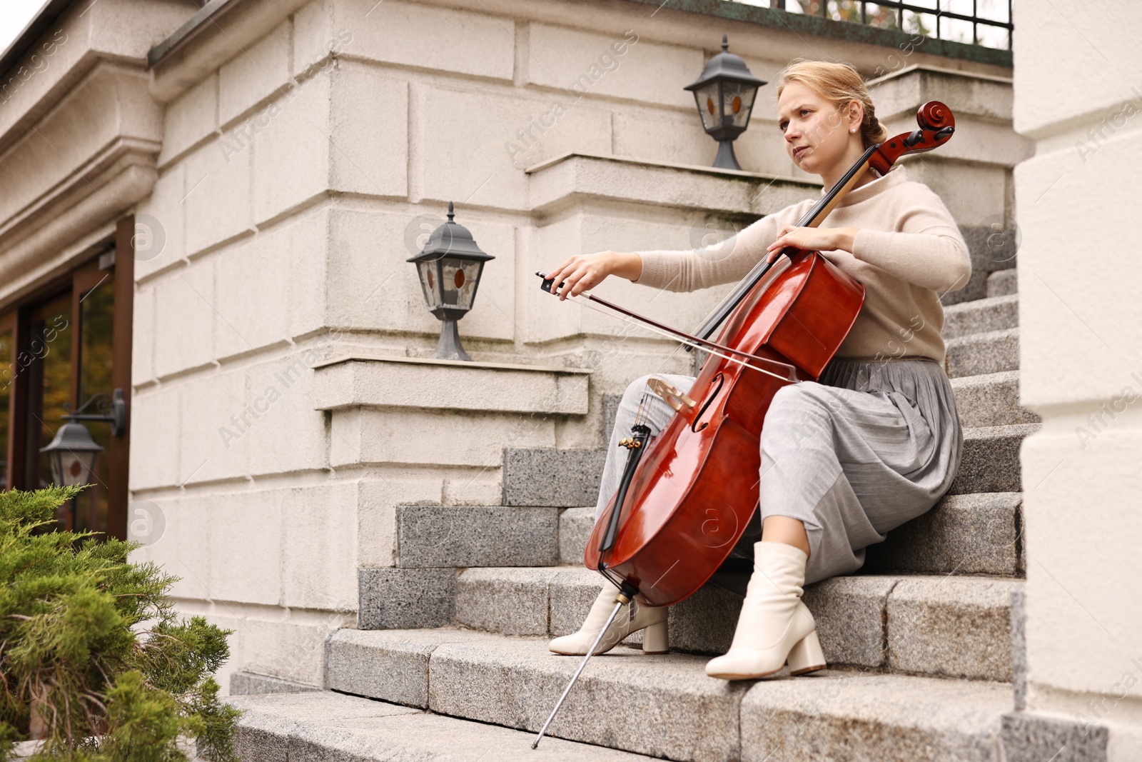 Photo of Beautiful young woman playing cello on stairs outdoors. Classic musical instrument