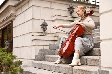 Photo of Beautiful young woman playing cello on stairs outdoors. Classic musical instrument