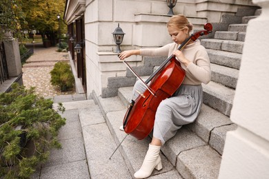 Beautiful young woman playing cello on stairs outdoors. Classic musical instrument