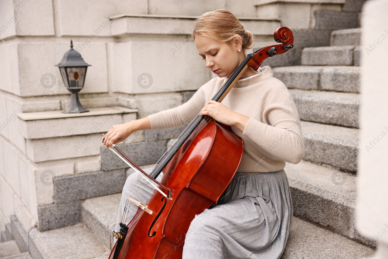 Photo of Beautiful young woman playing cello on stairs outdoors. Classic musical instrument