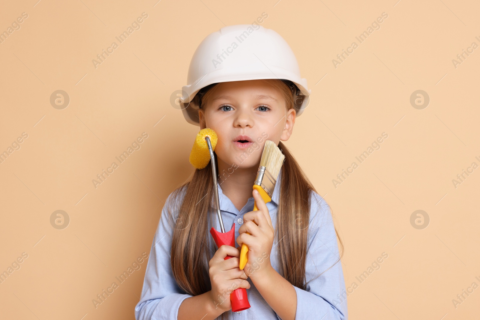 Photo of Little girl in hard hat with painting tools on beige background. Dreaming about future profession