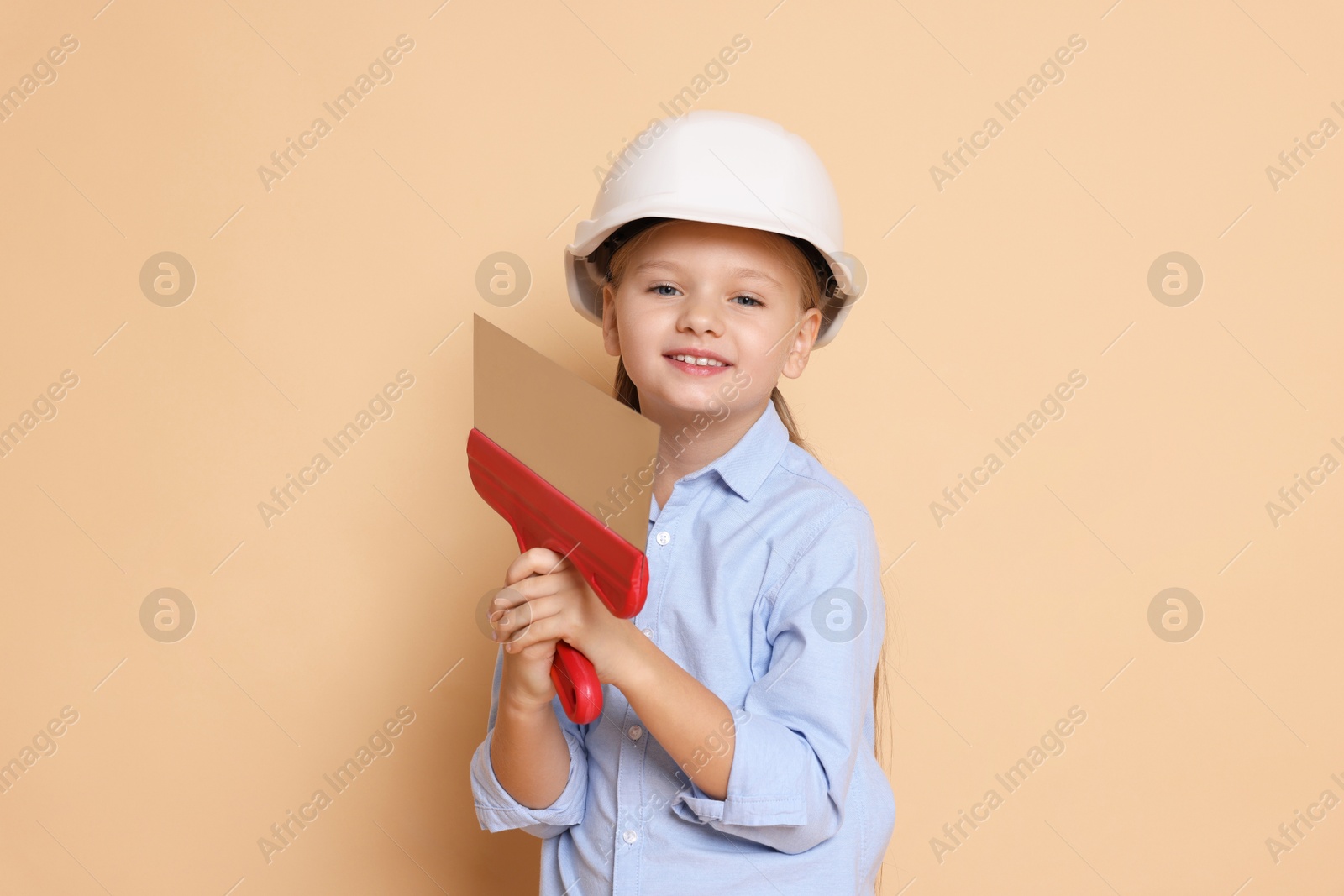 Photo of Little girl in hard hat with putty knife on beige background. Dreaming about future profession