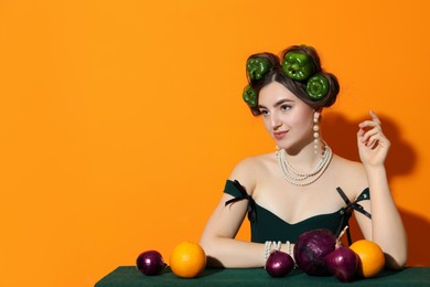 Photo of Young woman with peppers in her hair, fruits and vegetables at green table on orange background, space for text