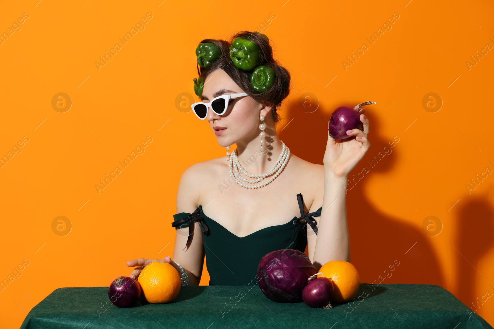 Photo of Young woman with peppers in her hair, sunglasses, fruits and vegetables at green table on orange background