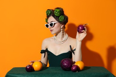 Photo of Young woman with peppers in her hair, sunglasses, fruits and vegetables at green table on orange background