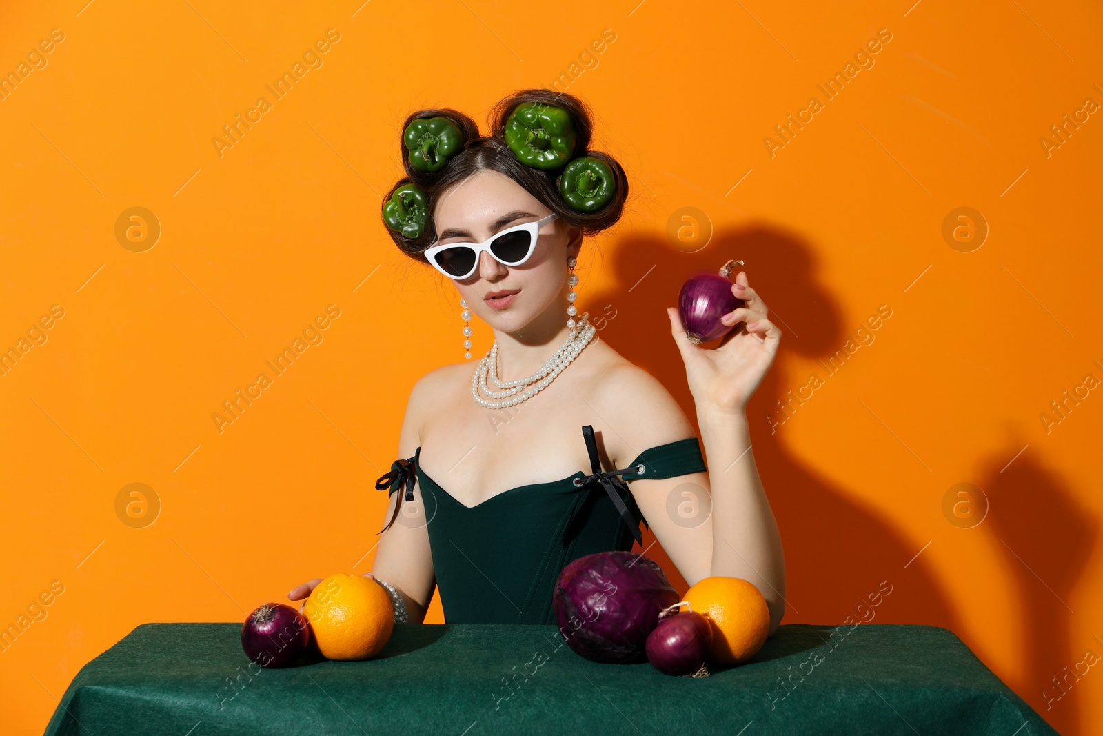 Photo of Young woman with peppers in her hair, sunglasses, fruits and vegetables at green table on orange background