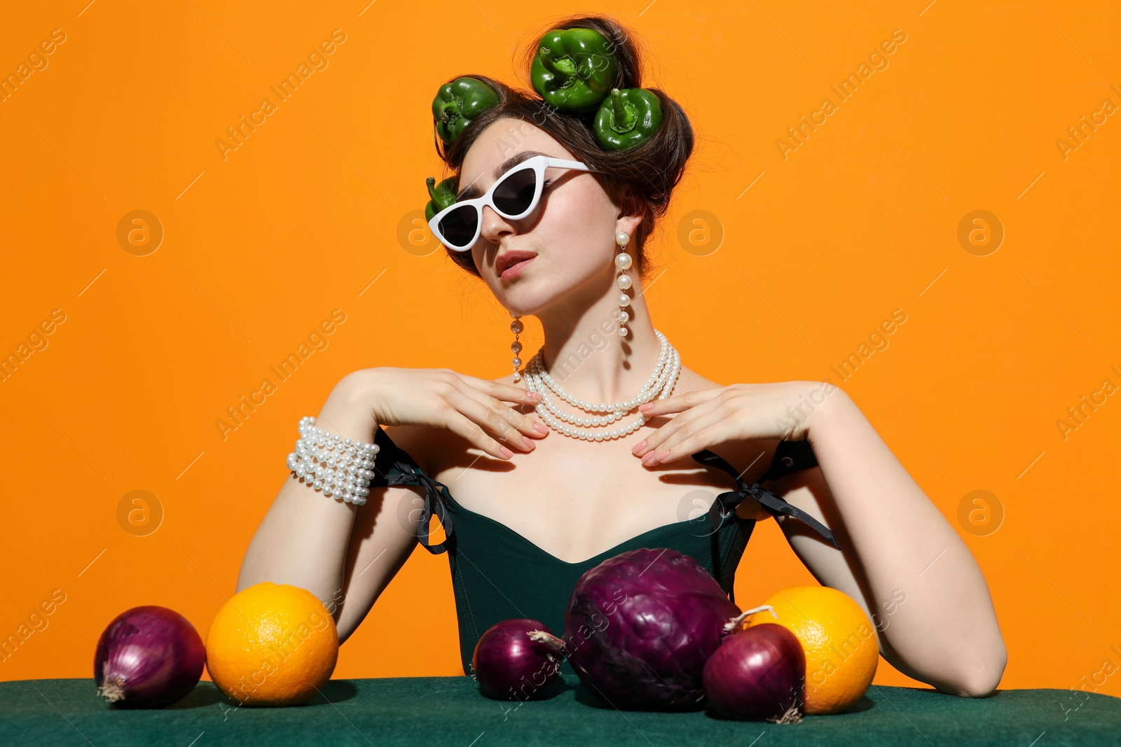 Photo of Young woman with peppers in her hair, sunglasses, fruits and vegetables at green table on orange background