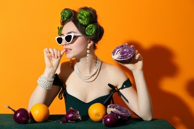 Photo of Young woman with peppers in her hair, sunglasses, fruits and vegetables at green table on orange background