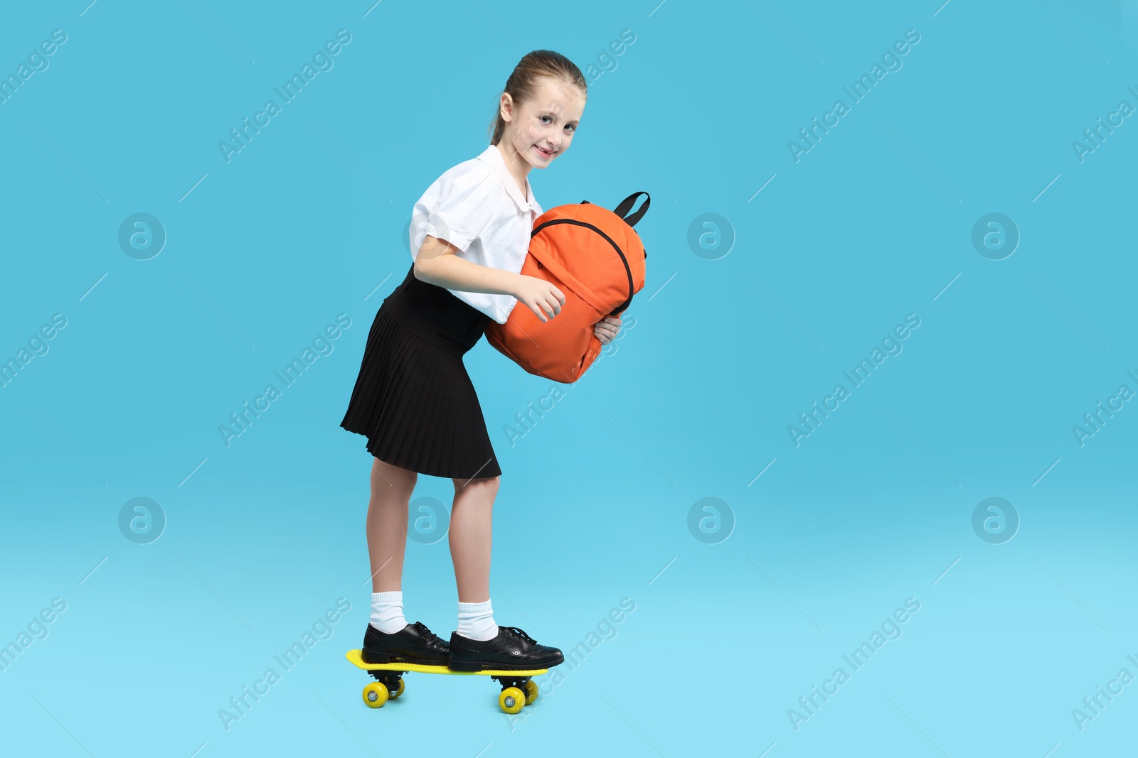 Photo of Little girl with backpack standing on penny board against light blue background