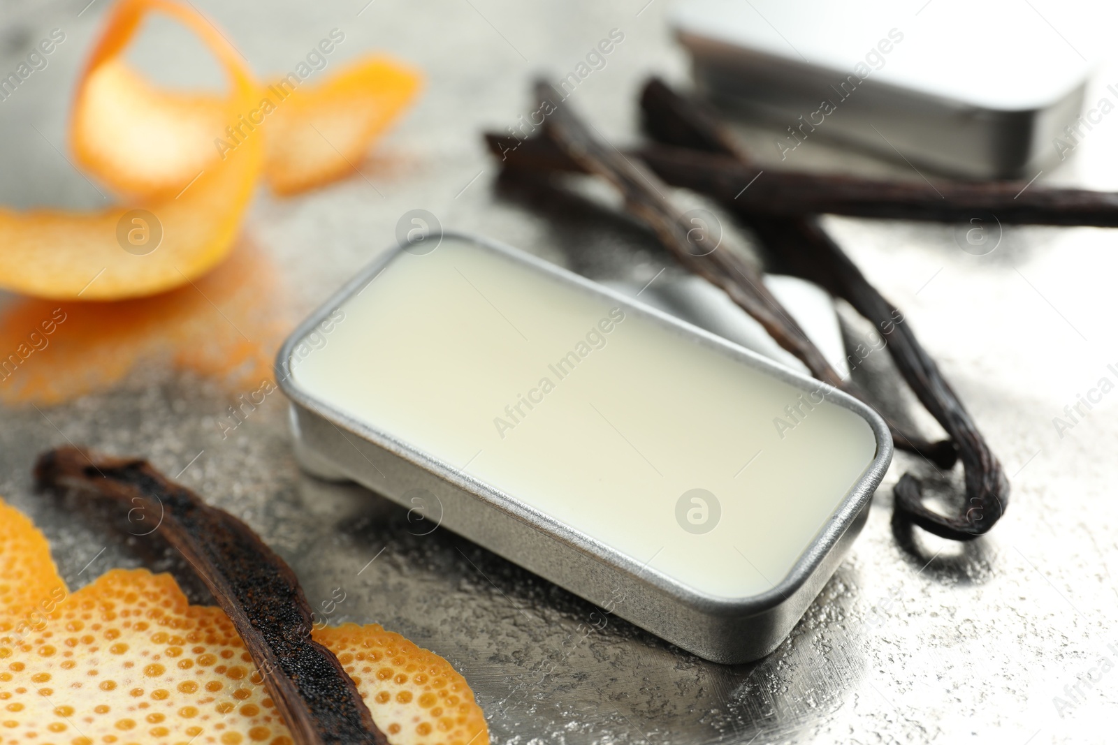 Photo of Natural solid perfume in container, vanilla pods and orange peels on grey table, closeup