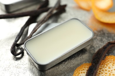 Photo of Natural solid perfume in container, vanilla pods and orange peels on grey table, closeup