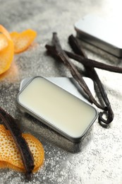 Photo of Natural solid perfume in container, vanilla pods and orange peels on grey table, closeup