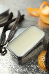 Photo of Natural solid perfume in container, vanilla pods and orange peels on grey table, closeup