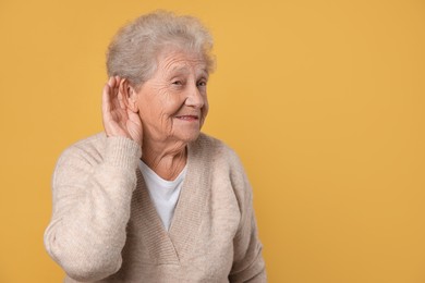 Photo of Senior woman showing hand to ear gesture on dark yellow background, space for text