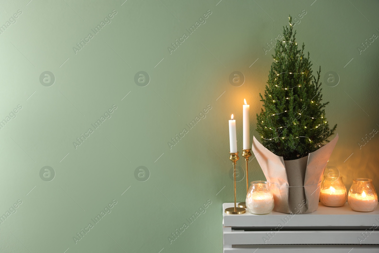 Photo of Small spruce tree decorated with Christmas lights and burning candles on white radiator cabinet near green wall, space for text