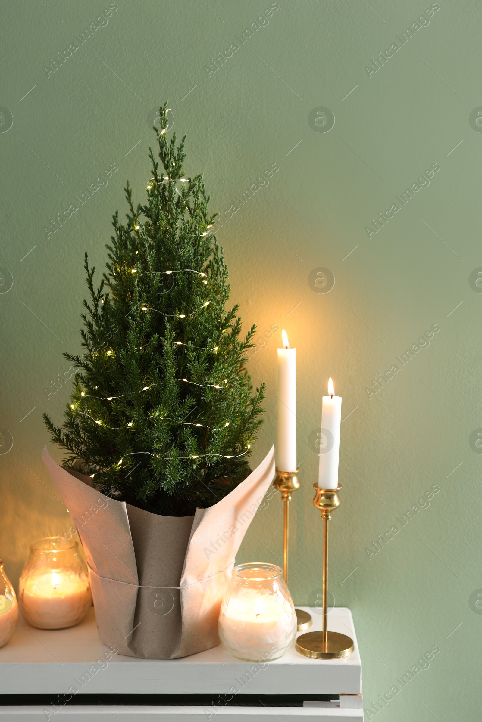 Photo of Small spruce tree decorated with Christmas lights and burning candles on white radiator cabinet near green wall