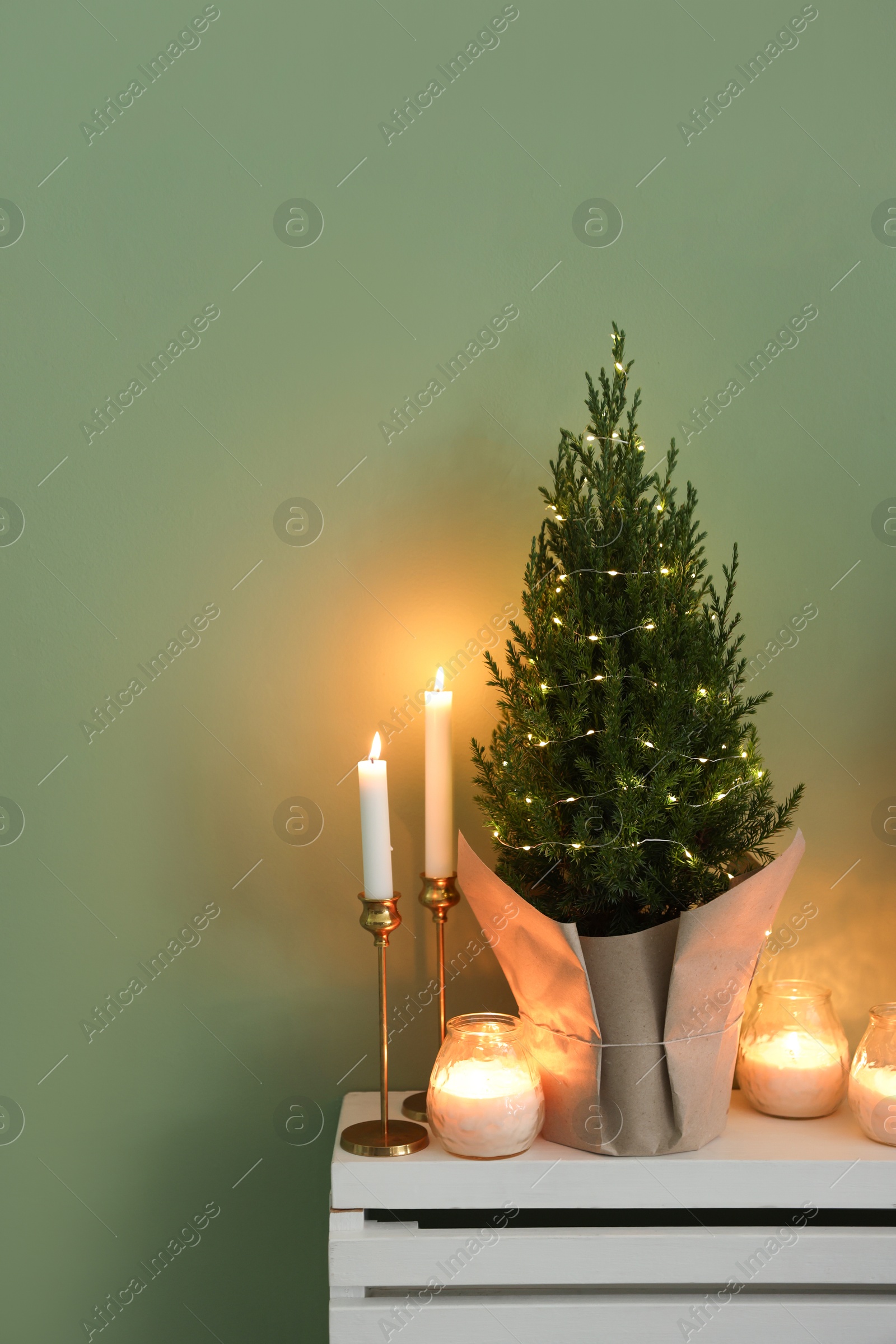 Photo of Small spruce tree decorated with Christmas lights and burning candles on white radiator cabinet near green wall