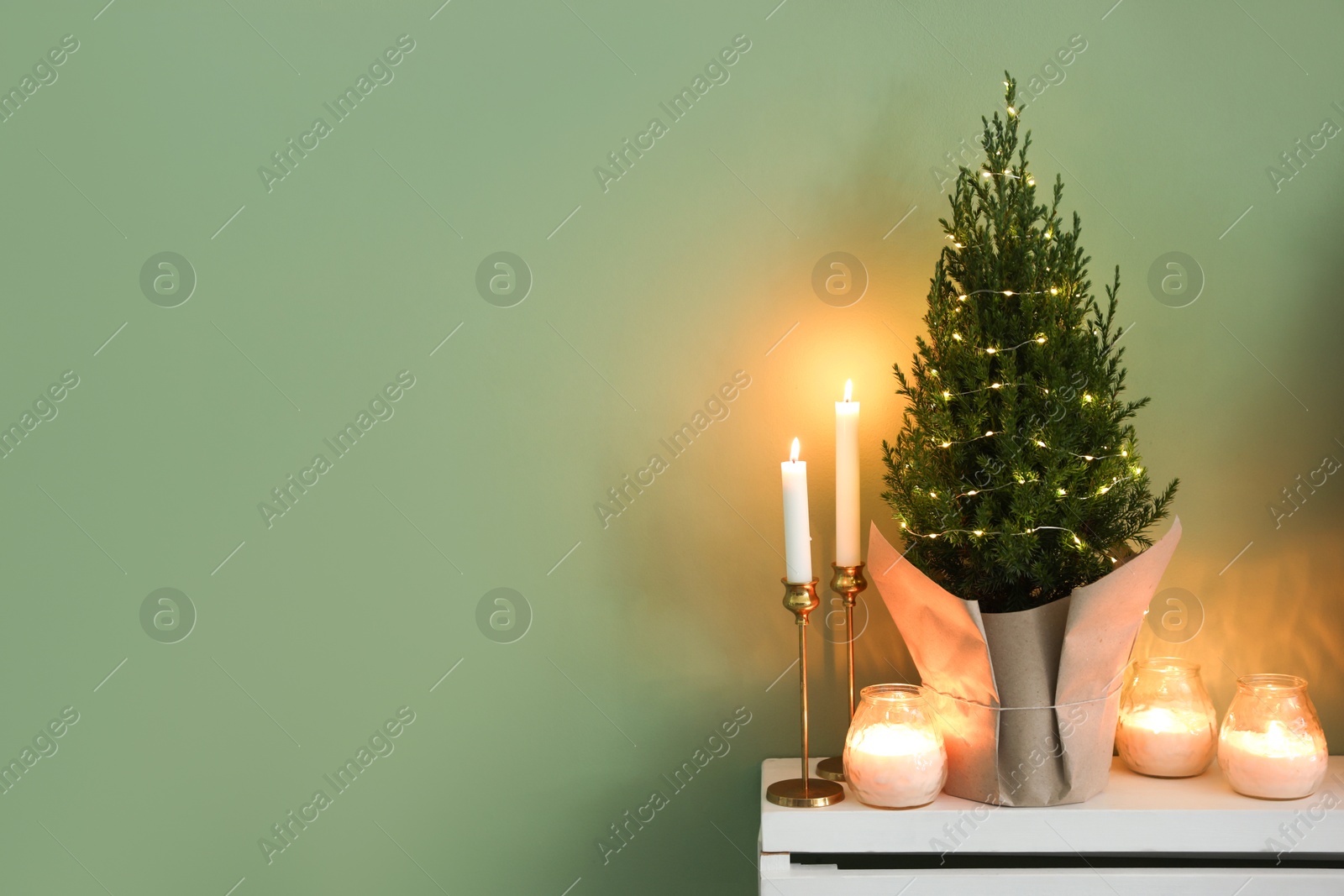 Photo of Small spruce tree decorated with Christmas lights and burning candles on white radiator cabinet near green wall, space for text