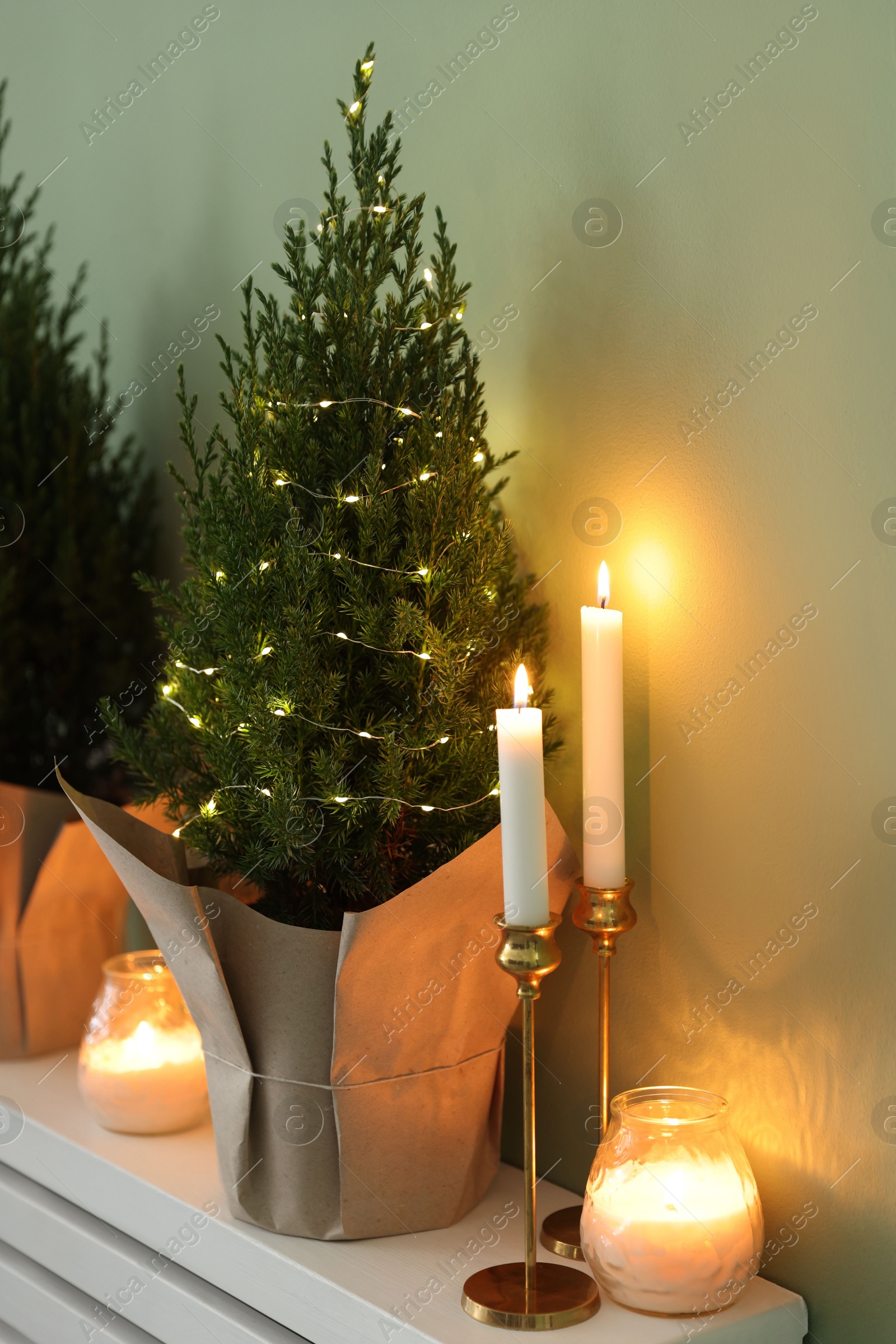 Photo of Small spruce tree decorated with Christmas lights and burning candles on white radiator cabinet near green wall