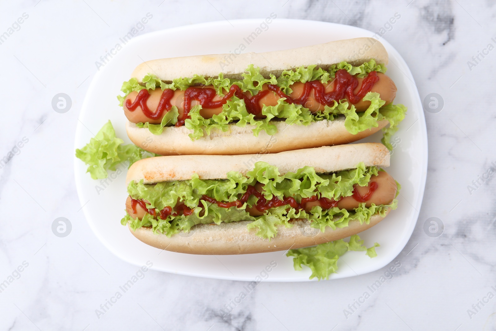 Photo of Tasty hot dogs with lettuce and ketchup on white marble table, top view