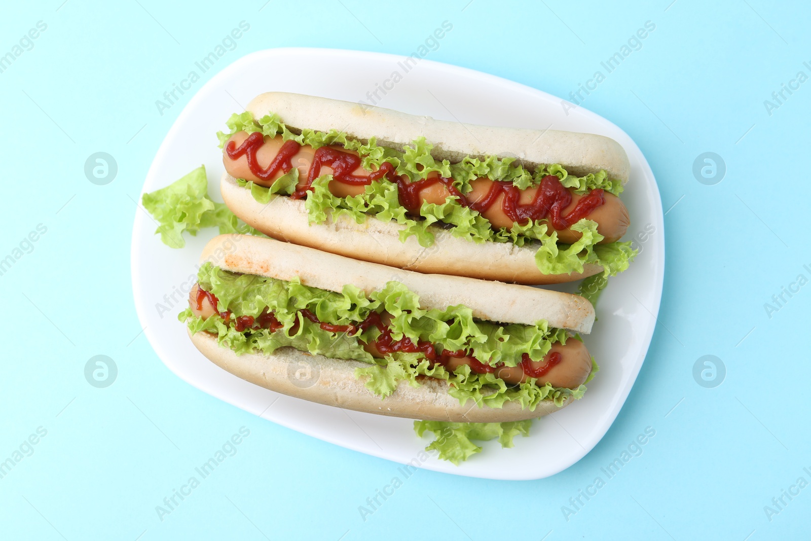 Photo of Tasty hot dogs with lettuce and ketchup on light blue background, top view