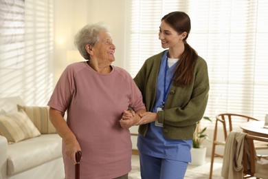 Photo of Caregiver assisting senior woman with walking cane indoors. Home health care service