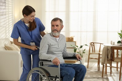 Photo of Caregiver assisting senior man in wheelchair indoors. Home health care service