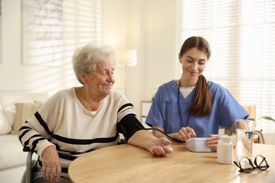 Photo of Caregiver measuring patient's blood pressure at table indoors. Home health care service