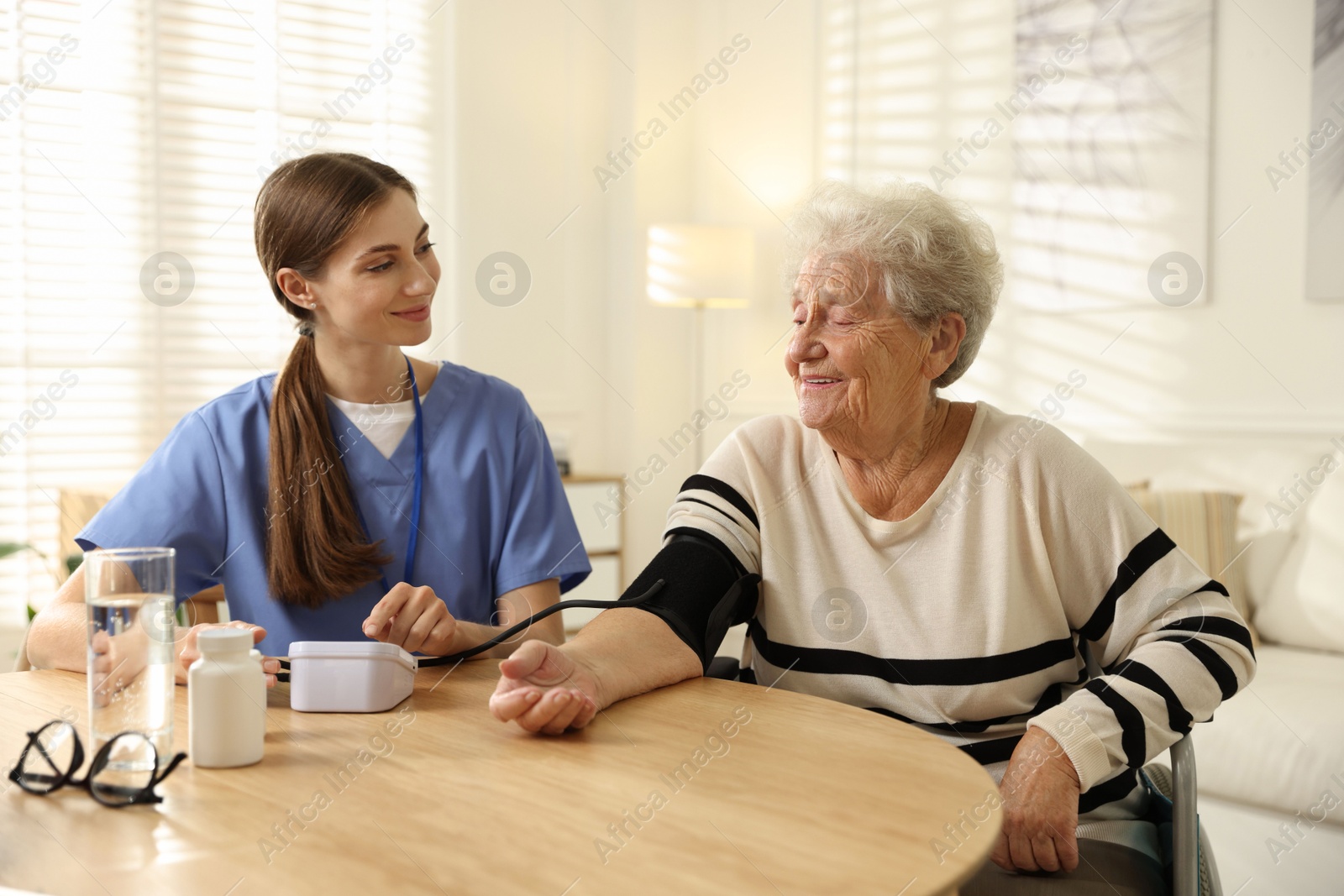 Photo of Caregiver measuring patient's blood pressure at table indoors. Home health care service