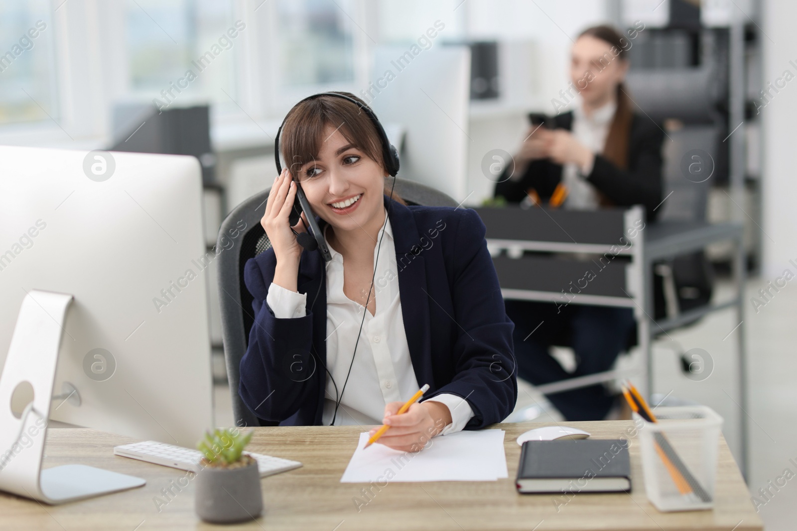 Photo of Saleswoman making notes while talking to client via headset at desk in office