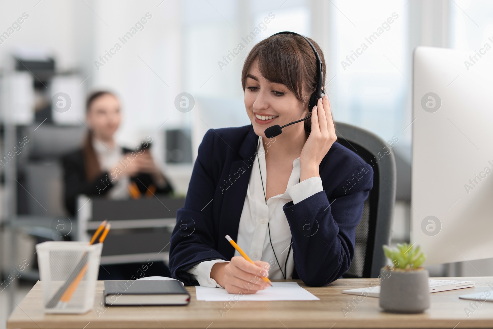 Photo of Saleswoman making notes while talking to client via headset at desk in office