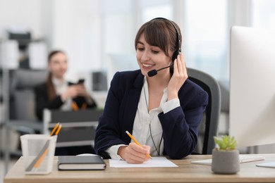 Photo of Saleswoman making notes while talking to client via headset at desk in office