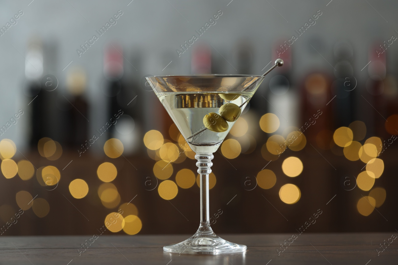 Photo of Martini cocktail with olives in glass on wooden table against blurred background. Bokeh effect