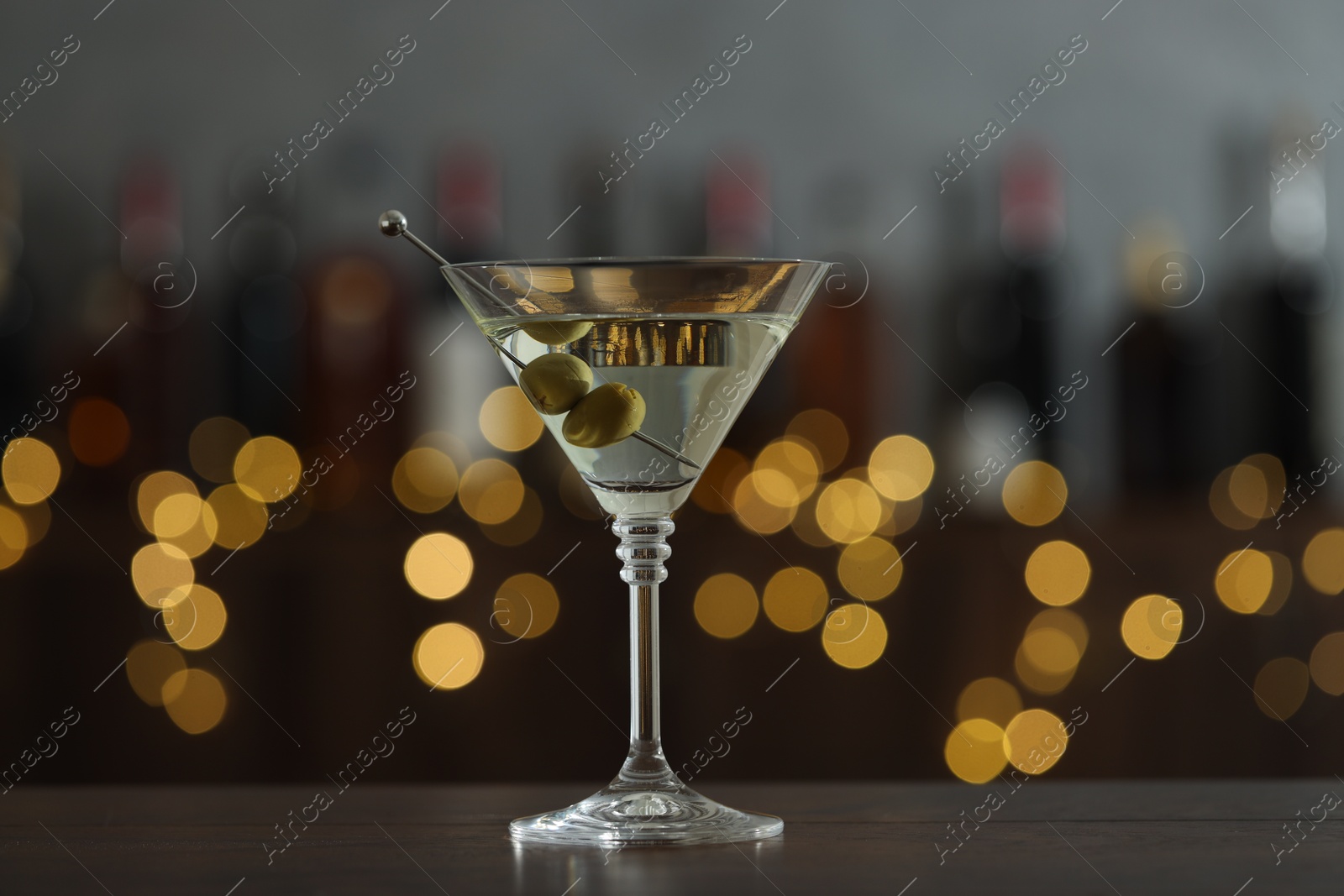 Photo of Martini cocktail with olives in glass on wooden table against blurred background. Bokeh effect