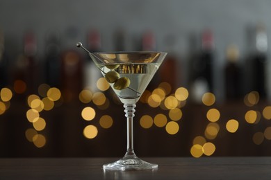 Photo of Martini cocktail with olives in glass on wooden table against blurred background. Bokeh effect