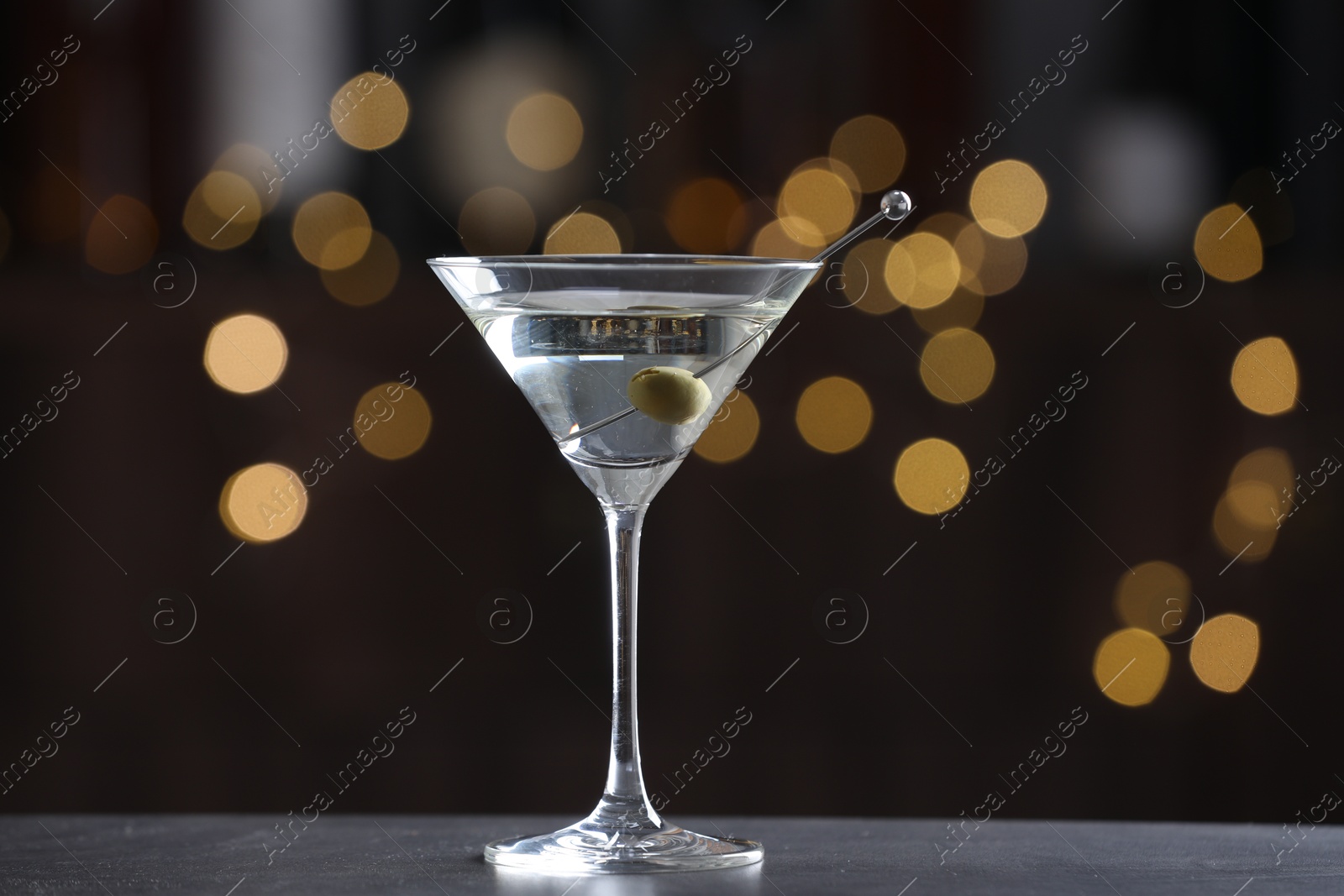 Photo of Martini cocktail with olive in glass on grey table against blurred background. Bokeh effect