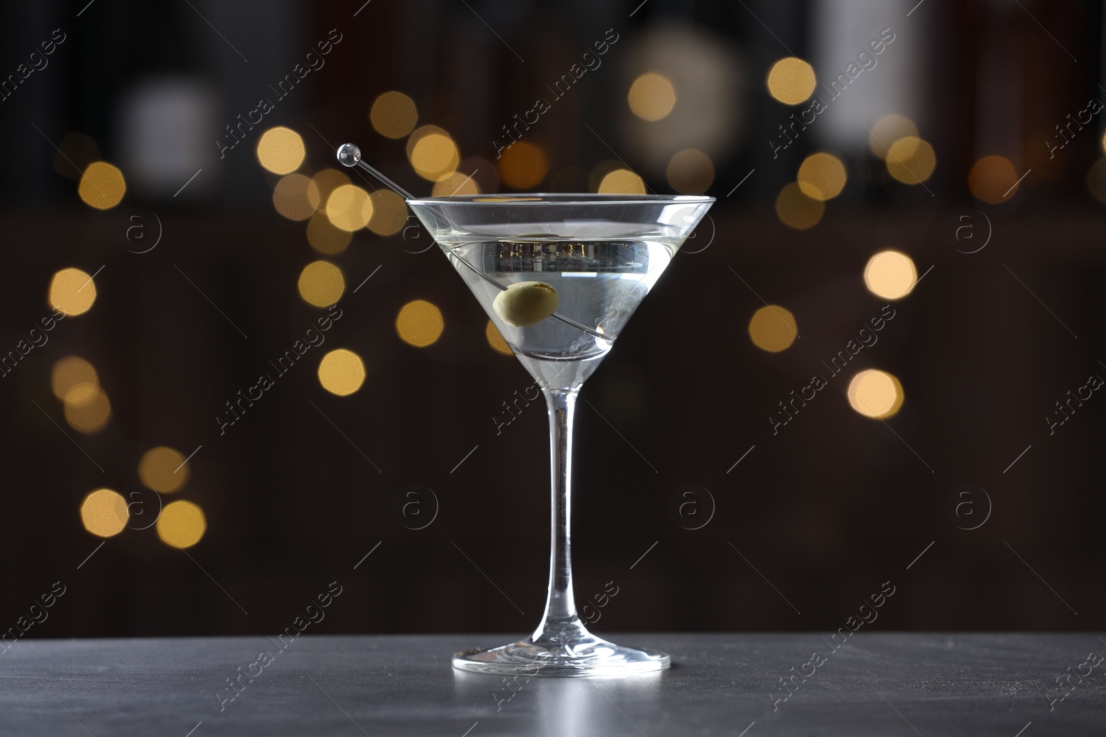 Photo of Martini cocktail with olive in glass on grey table against blurred background. Bokeh effect