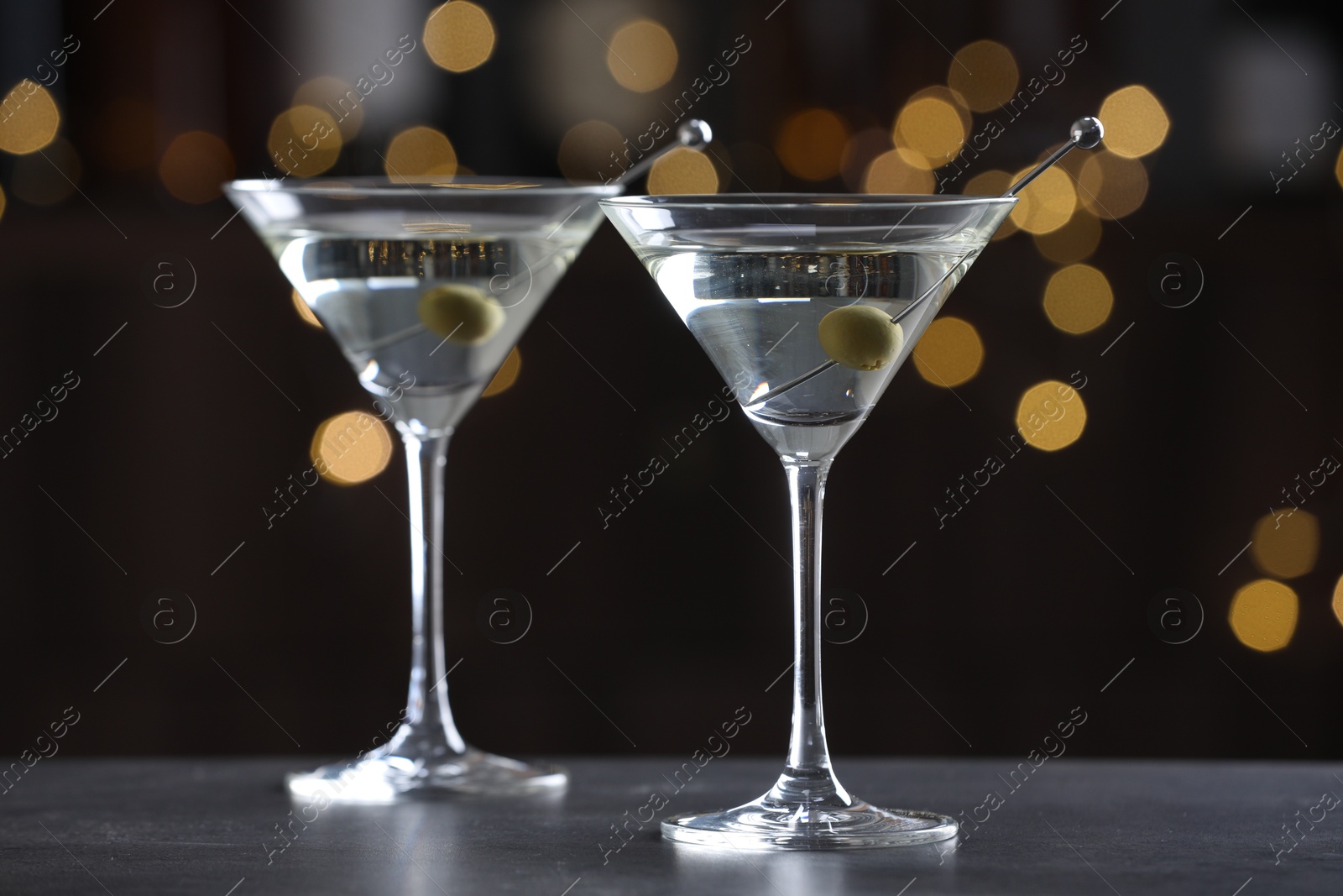 Photo of Martini cocktail with olives in glasses on grey table against blurred background, closeup. Bokeh effect