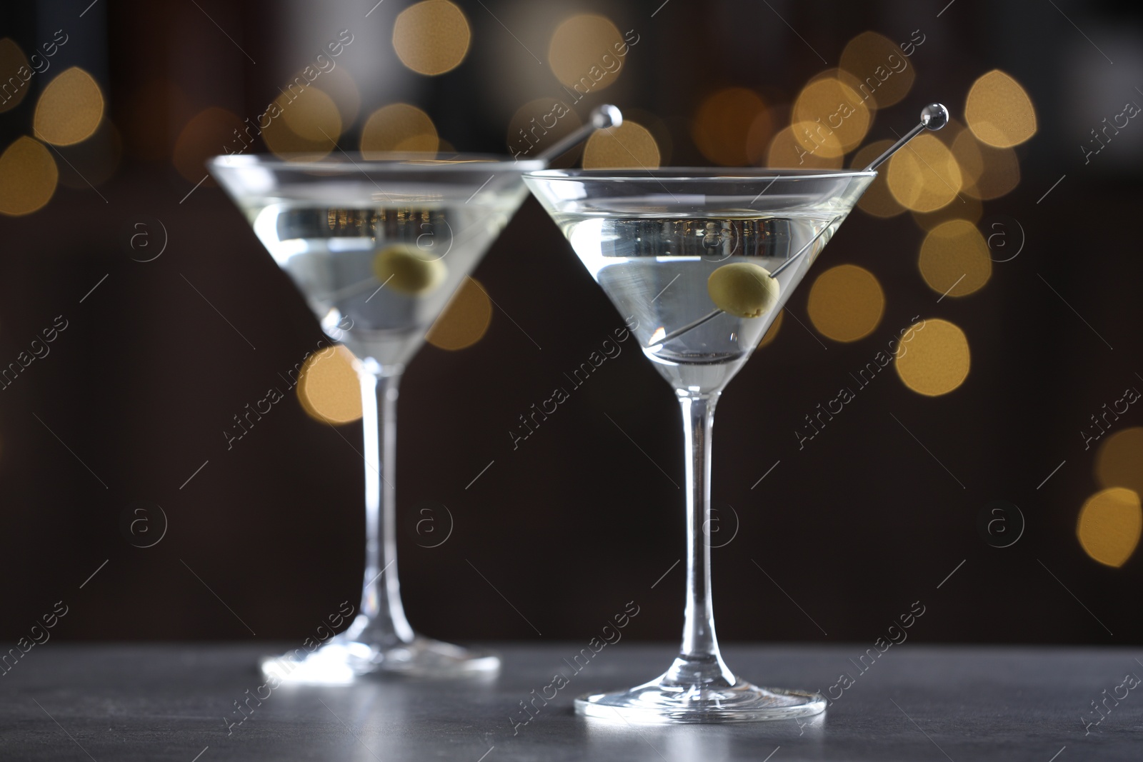 Photo of Martini cocktail with olives in glasses on grey table against blurred background, closeup. Bokeh effect