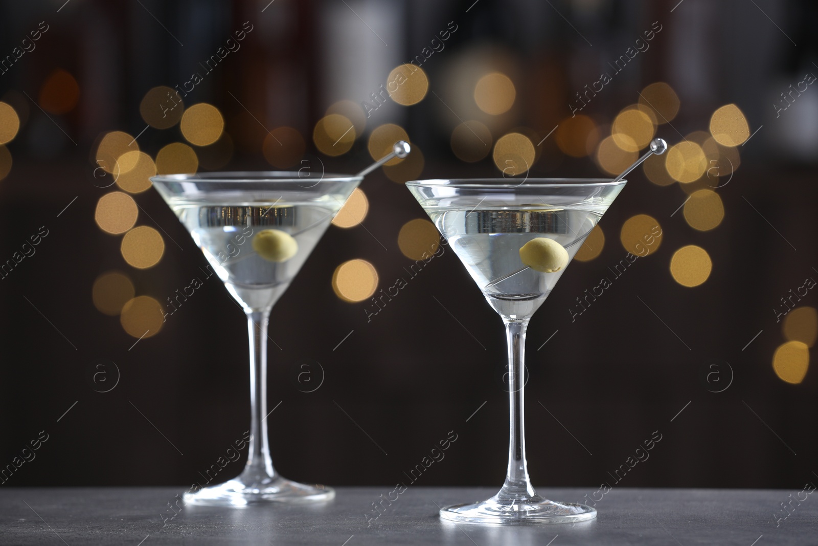 Photo of Martini cocktail with olives in glasses on grey table against blurred background. Bokeh effect